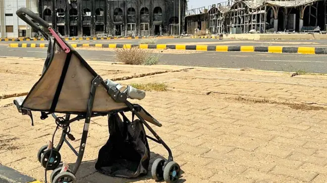 A pram abandoned on a pavement outside Khartoum's airport. The blackened shells of the terminal buildings can be seen behind - March 2025.