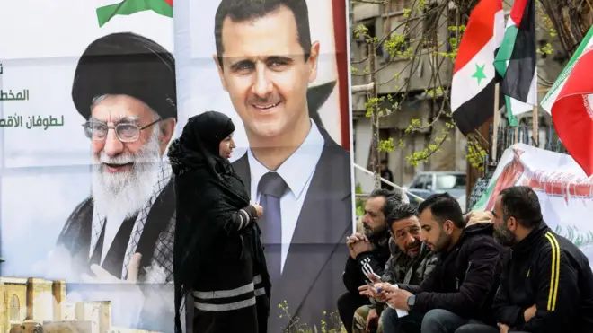 پرتره‌هایی از بشار اسد، رئیس‌جمهور سابق سوریه (راست)، و آیت‌الله علی خامنه‌ای، رهبر جمهوری اسلامی ایران، در حالی دیده می‌شوند که گروهی از فلسطینی‌ها در ورودی اردوگاه یرموک برای پناهندگان فلسطینی در جنوب دمشق نشسته‌اند. این عکس در جریان توزیع کمک‌های بشردوستانه‌ از سوی ایران و به مناسبت ماه رمضان، در تاریخ ۲۶ مارس ۲۰۲۴ گرفته شده است. (عکس از لؤی بشاره / خبرگزاری فرانسه)