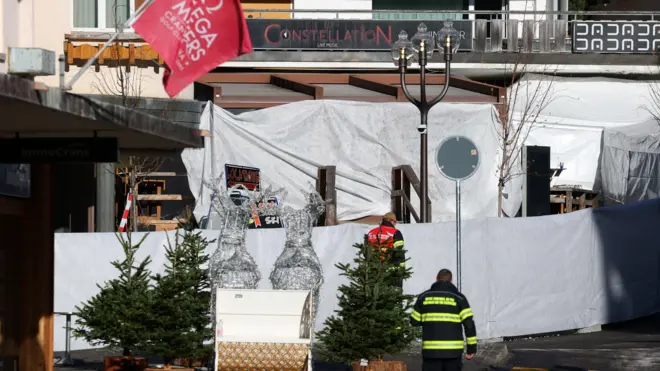 A wide shot shows the front of Le Constellation bar covered in white tarpaulin, with an emergency worker standing in the foreground. Christmas trees and model reindeer are also visible in front of the bar. 
