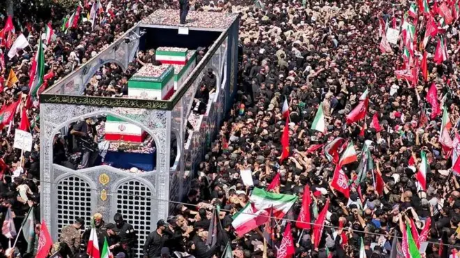 د زرګونه خلکو ترمنځ یو لوی موټر روان برېښي چې تابوتونه پکې د ایران په بیرغ کې پوښل شوي ایښي دي.