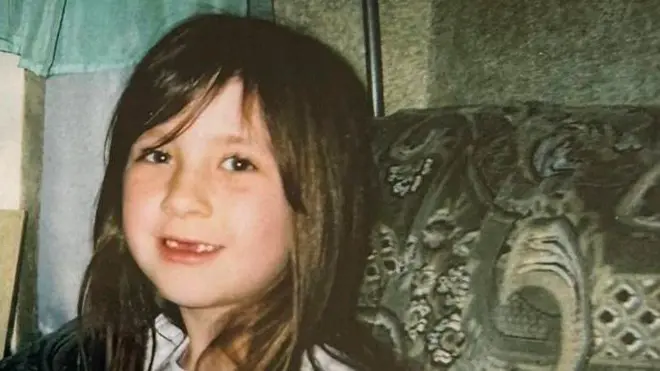 Yaklaşık altı yaşında olan küçük Gabby'nin, yeşil bir kanepede otururken kameraya gülümsediği bir aile fotoğrafı. Uzun kahverengi saçları, perçemi ve ön süt dişlerinin dökülmesinden dolayı ağzının üst kısmında oluşan boşluk var.