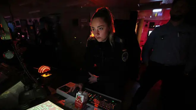 Bir kadın denizaltı subayı kızıl karanlık bir ortamda ileriye bakıyor, bir takım tuşlara dokunuyor