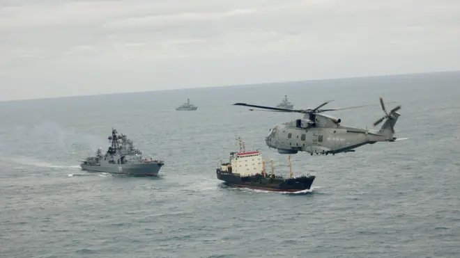 Над морем летить військовий вертоліт, нижче в воді рухаються кілька кораблів, серед них військові судна та одне цивільне. Море спокійне, небо хмарне; сцена виглядає як військове патрулювання або супровід у відкритому морі.