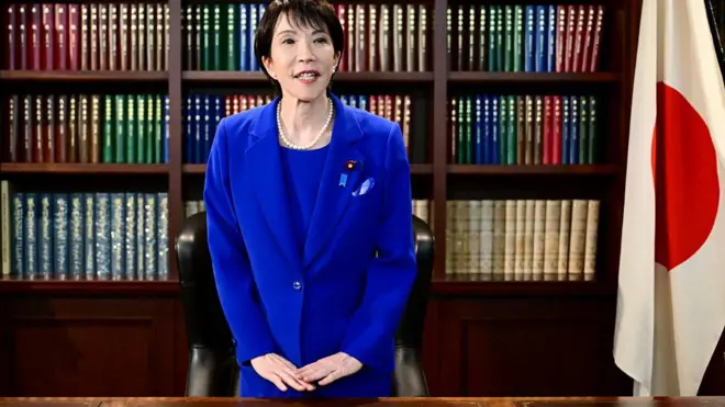 Sanae Takaichi em pé, falando, diante de mesa de escritório e ao lado de bandeira do Japão