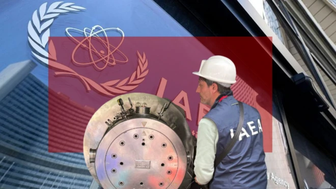 Человек в белой защитной каске и синем жилете с белыми буквами IAEA («МАГАТЭ») на спине. Он стоит рядом с металлическим цилиндром.