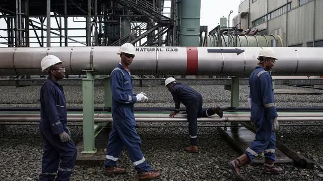 Gas workers dress for protective gear waka past large pipeline labelled 'natural gas'.