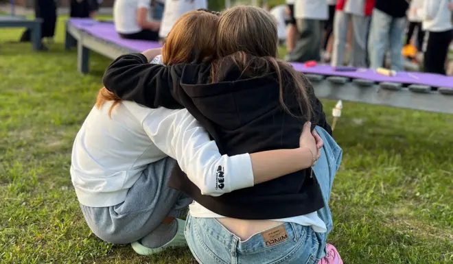 Two teenage girls sit with their arms around each other, hugging, while sitting in front of a camp fire