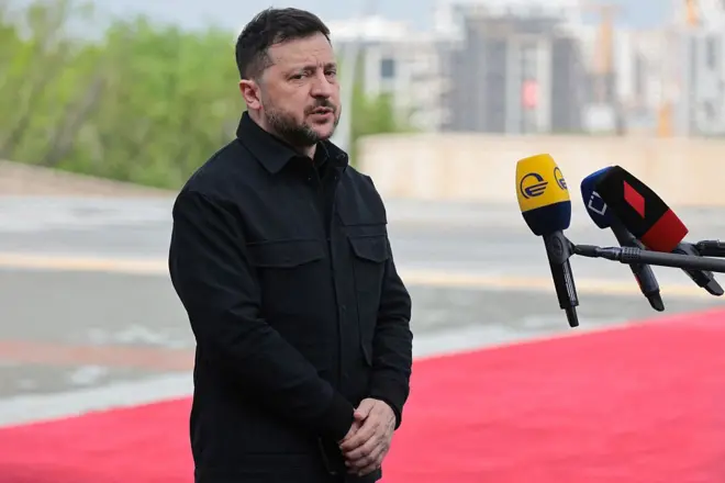 Зеленський у чорному стоїть на червоній доріжці перед мікрофонами 