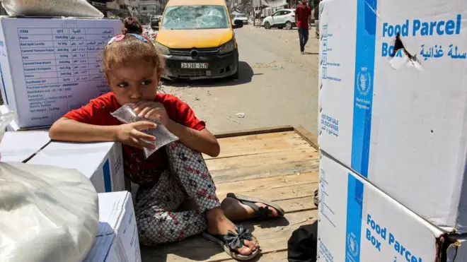 Una niña sostiene una bolsa de plástico transparente llena de agua mientras se sienta en un carro junto a los paquetes de ayuda humanitaria.