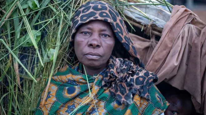 Neema akiwa mbele ya kibanda anakoishi, nyuma yake kuna mtoto anayetazama kamera wakati picha inapigwa.