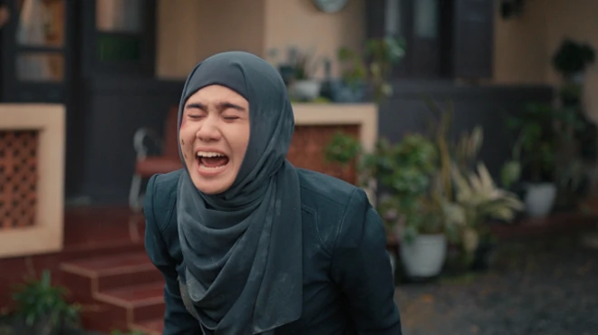 A still from the movie, which shows Norma in dark blue tudong screaming outside a house.
