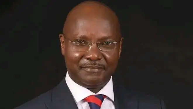 Man wey dey look di camera, e wear glass, white shirt, black suit and bald head