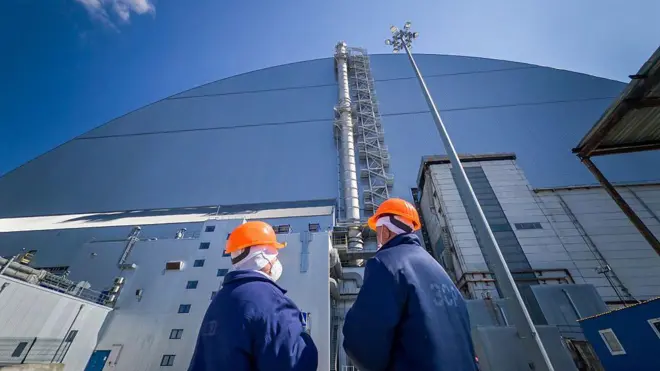 Двое работников ЧАЭС в оранжевых шлемах стоят у купола, защищающего саркофаг