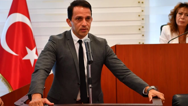 Mücahit Birinci kürsüde konuşuyor, arkasında Türk bayrağı ve oturan bir kadın