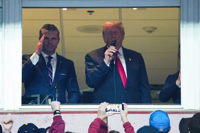 Pete Hegseth faz saudação com mão na cabeça, denotando obediência, ao lado de Trump, que fala no microfone em camarote, assistido por plateia que tira fotos