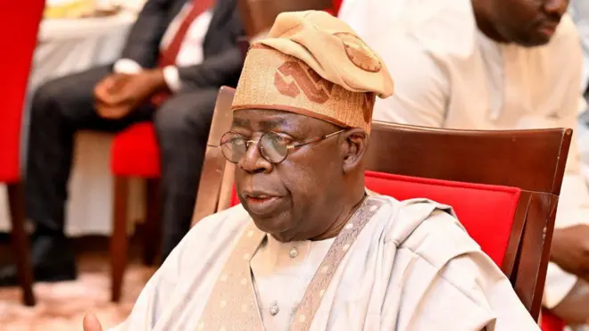 Nigeria President, Bola Tinubu wear cream colour cloth wit brown designs e also wear cream colour cap with broun design and eye glasses as e siddon for wooden chair with red design.