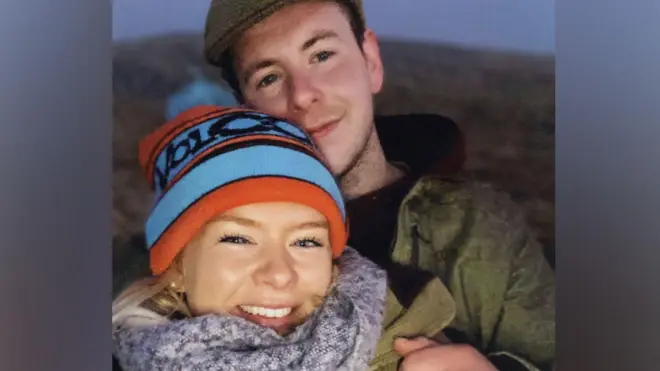 A médica Caroline Muirhead, usando um gorro de lã azul e vermelho e um cachecol cinza, com Alexander “Sandy” McKellar ao fundo, de boné e casaco escuro