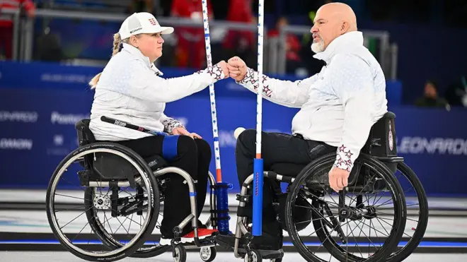 Американские керлингисты Лора Дуайер и Стивен Эмт сидят на колясках и протягивают друг другу руки в одобряющем жесте