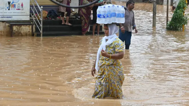 వరద నీటిలో నీళ్ల బాటిళ్లు మోసుకెళ్తున్న మహిళ