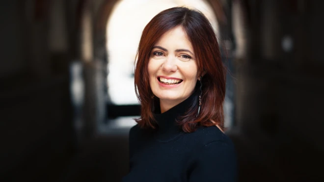 Ilana Eleá em uma foto olhando para a câmera e sorrindo. Ela tem cabelos escuros, lisos, na altura dos ombros e veste uma blusa escura.