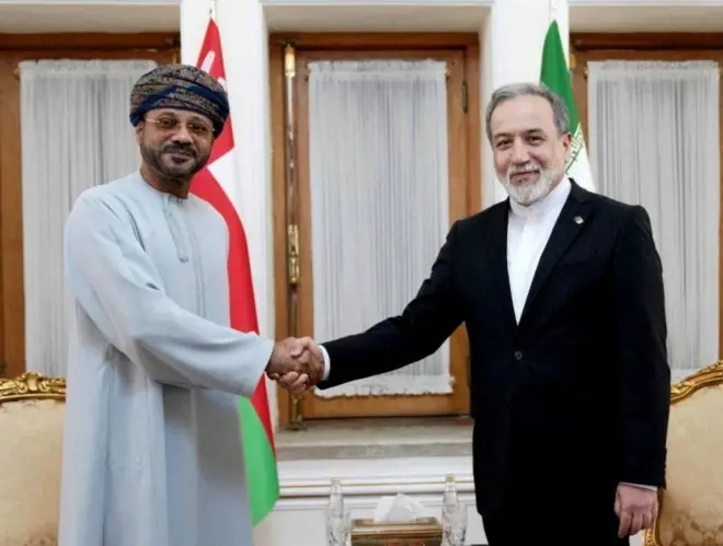 عراقجي يرتدي بدلة رسمية سوداء مع قميص أبيض دون ربطة عنق، وبجانبه البوسعيدي يرتدي ثوباً تقليدياً أبيض اللون وخلفهما علما بلديهما.