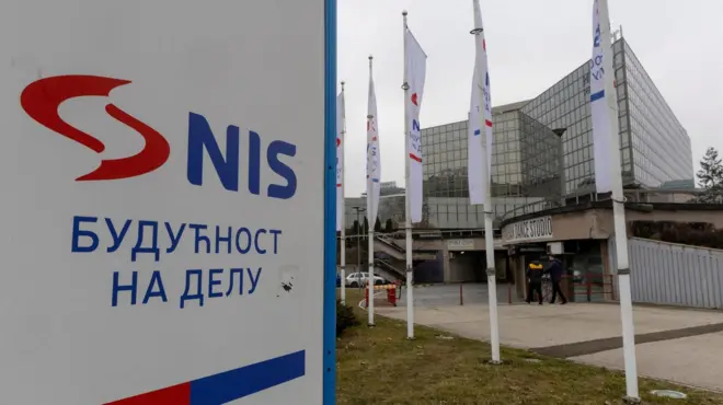 Velika tabla na kojoj je napisano plavim slovima NIS, crveni znak firme pored, a ispod ćirilično - Budućnost na delu. U daljini je staklena zgrada kompanije. 