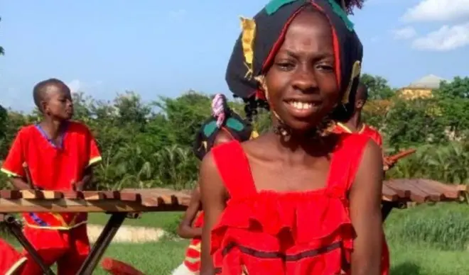 One girl and one boy wear red cloth dem dey play local Xylophone