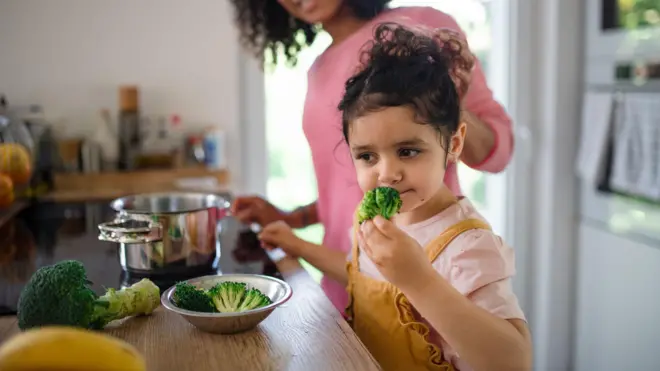 طفلة تقف في المطبخ أمام طاولة تشتم البروكلي