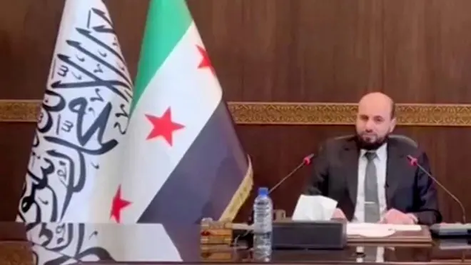 Man in suit sits at head of a table with big flags behind him
