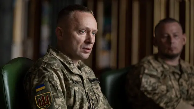 Близько 40-річний чоловік у військовій формі Збройних сил України сидить за столом під час наради. На рукаві видно український прапор і шеврон із тризубом. На задньому плані, розмитий, сидить Павло Паліса.