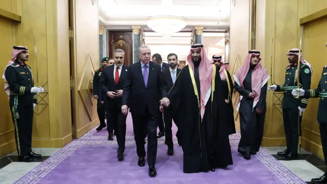 Cumhurbaşkanı Erdoğan, Suudi yerel kıyafeti giyen Prens ile yan yana yürüyor. Bir holde ilerliyorlar ve askerler kılıçla selam duruyor