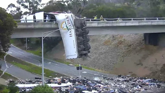 A truck hangs off an overpass bridge with its contents spilled across the motorway below