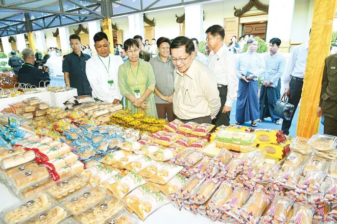 ပေါင်မုန့် ကိတ်မုန့်တွေ ဖြန့်ခင်းထားတဲ့ စားပွဲကို လက်အနောက်ပစ်ပြီး ငုံ့ကြည့်နေတဲ့ ဦးမင်းအောင်လှိုင်နဲ့ ဘေးနဲ့အနောက်မှာ သူ့ကိုရပ်ကြည့်နေတဲ့ သူတွေ 