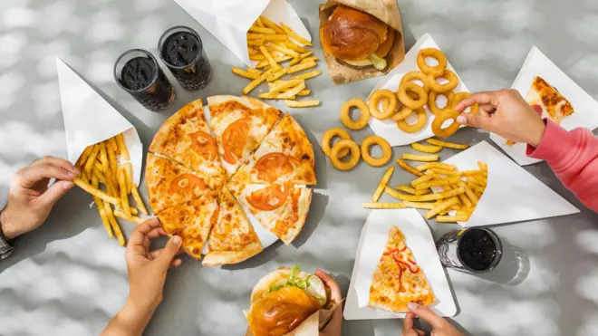 Pizza tabakları, hamburger ekmekleri, soğan halkaları, patates kızartması ve kolalı içeceklerle dolu bir masa. Hepsi ultra işlenmiş gıdaların örnekleri. Eller uzanıp farklı yiyecekleri alıyor.

