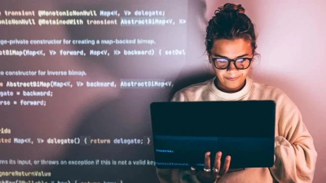 Стоковое фото, изображающее программистку за работой. Она держит в руках ноутбук, а рядом на фоне строчки кода.