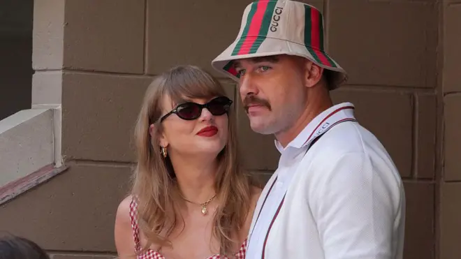 Taylor Swift, wearing a red gingham top, looking up at Travis Kelce, wearing a striped Gucci bucket hat