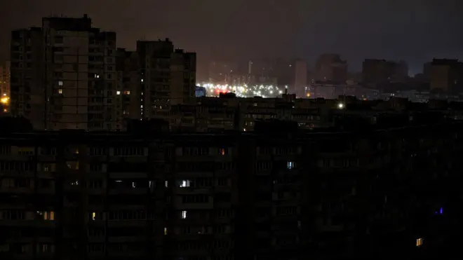 Блекаут у спальному районі Києва: лише в поодиноких вікнах багатоповерхівок видно світло. За ними – яскраво освітлена ліхтарями вулиця.