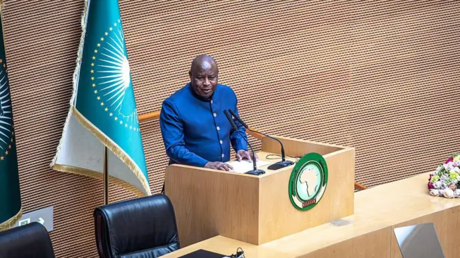 Perezida w'u Burundi Évariste Ndayishimiye arimo kuvuga ijambo mu ndangururamajwi ebyiri, mu cyumba cy'inama cyitwa 'Nelson Mandela Hall', mu nama isanzwe ya 39 y'inteko y'umuryango w'Ubumwe bw'Afurika (AU), ku cyicaro cy'uwo muryango i Addis Ababa muri Ethiopia, ku wa gatandatu, ku itariki ya 14 Gashyantare (2) mu mwaka wa 2026. Yambaye ikoti ry'ubururu ry'amaboko maremare n'ikora rigufi.