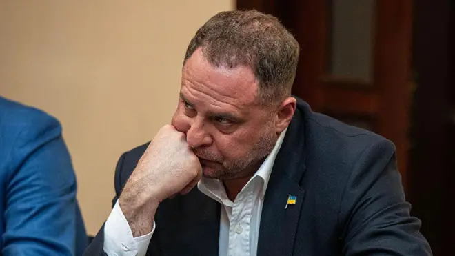 Крупний план: Єрмак в темному піджаку й білій сорочці сидить за столом, спираючи підборіддя на руку. На піджаку — маленький значок із прапором України.