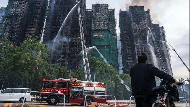 Awọn panapana n na ọpa omi si oke fiofio, sara awọn ọpọ ileesẹ alaja pupọ ti ina alatagba n jo nilu Hongkong, ọkọ ileesẹ panapana kan wa nilẹ, ti ọkunrin kan to wọ asọ kootu dudu si duro, o fa ọkada kan lọwọ, o n wo ile to n jo naa.