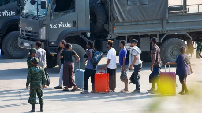 የካቲት ላይ የተለቀቁ ኢትዮጵያውያን በሰልፍ ሻንጣቸውን እየገፉ፤ ከፊታቸው ሁለት የታይላንድ ወታደሮች ይታያሉ