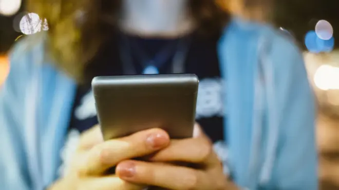 Obinrin to wọ aṣọ aláwọ̀ búlúù mu foonu lọwọ, tó sì jókòó sórí àga kan 