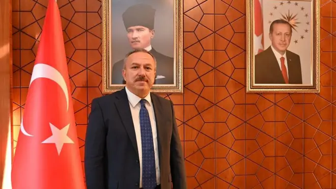 Ali Fidan lacivert bir takım elbise, beyaz gömlek ve mavi bir kravat takmış. Solunda Türk bayrağı duruyor, arkasındaki duvarda Atatürk portresi sağ tarafta da Erdoğan portresi asılı.