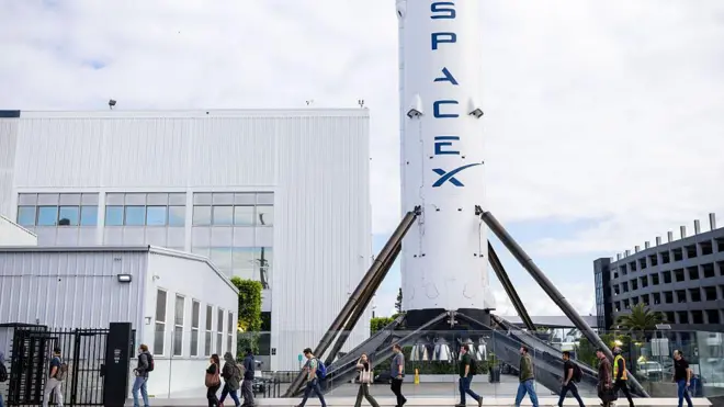 Uma fila de pessoas passa por um grande foguete branco com o logotipo azul Space X escrito nele.