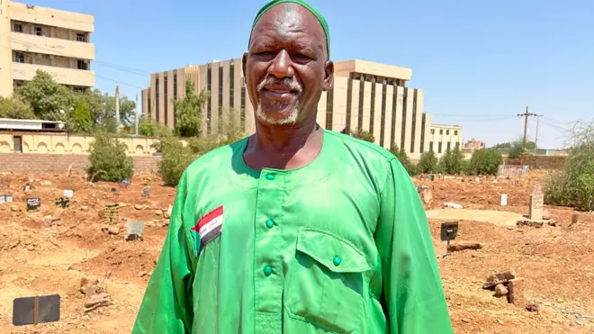 Abidin Durma, fossoyeur d'Omdurman, se tient dans le cimetière d'Ahmed Sharfi. Il porte une dishdasha, ou khaftan, vert vif, sur laquelle est cousu le drapeau du Soudan, ainsi qu'un chapeau kufi de la même couleur.