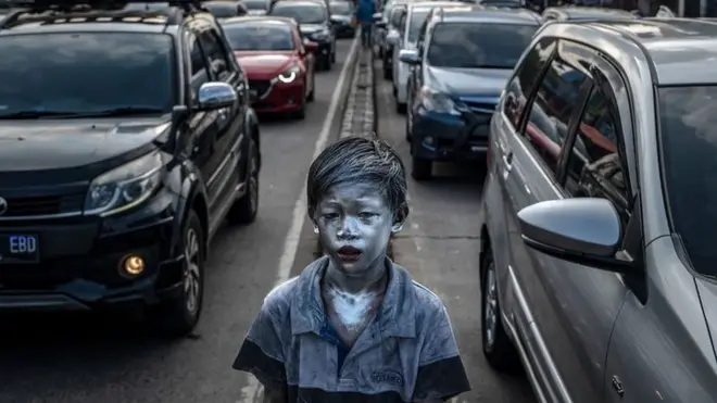 A boy whose skin appears metallic standing next to cars in traffic