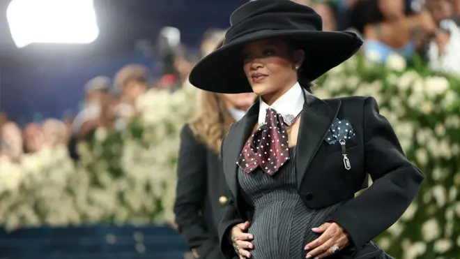 Rihanna appears at the 2025 Met Gala visibly pregnant. She is wearing a black and grey tailored outfit and large brim hat.