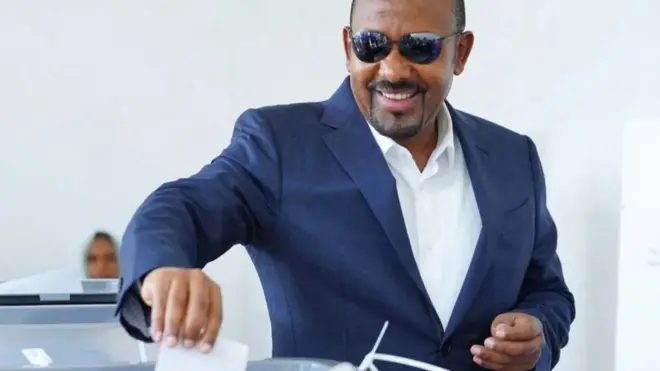 Abiy Ahmed yambaye ikositime y'ubururu, ishati yera, n'amarori y’izuba, ariko aramwenyura mu gihe ariko ashira ikarata y’itora mw’isandugu
