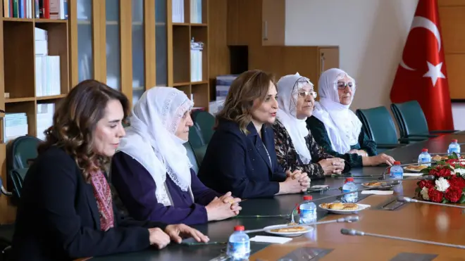 beyaz tülbentli Barış Anneleri DEM Parti sözcüsü Ayşegül Doğan'ın yanında bir masada oturuyor ve kenarda türk bayrağı var