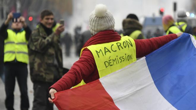 一名穿上寫有「馬克龍辭職」字樣的女示威者手持法國國旗在巴黎凱旋門前走過（24/11/2018）